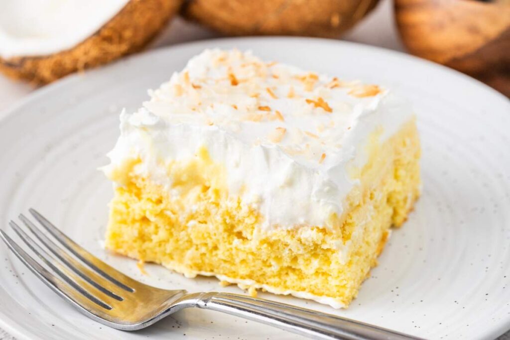 A slice of coconut cream poke cake topped with white frosting and sprinkled with toasted coconut served on a white plate with a fork beside it.