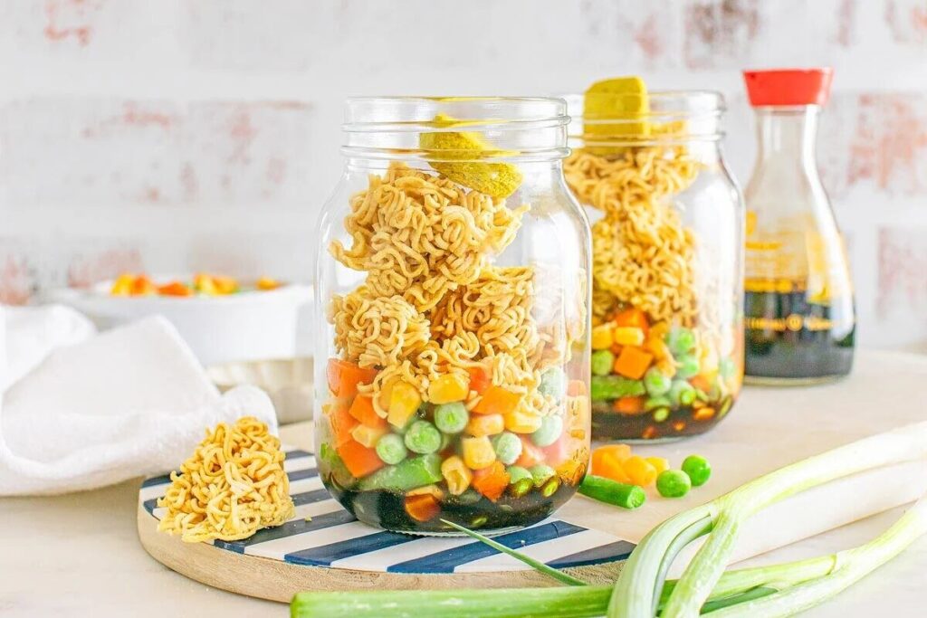 Two mason jars filled with layers of peas, corn, carrots, and dry ramen noodles sit on a striped board.