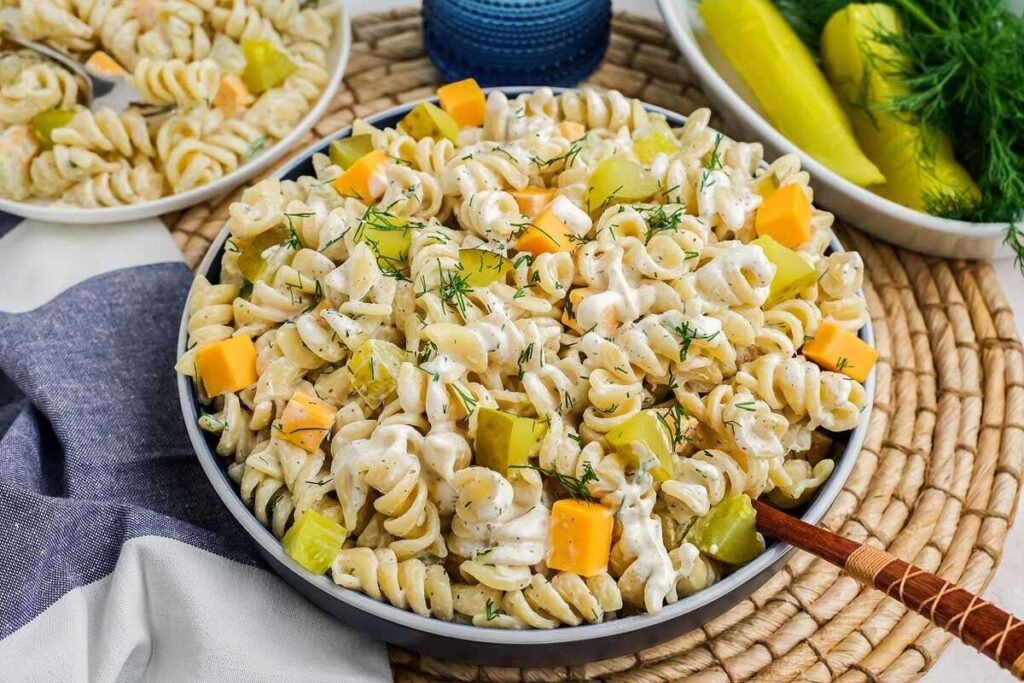 A bowl of dill pickle pasta salad with cheese cubes, rotini, and fresh dill sits on a woven mat beside a blue cloth and a bowl of pickles.