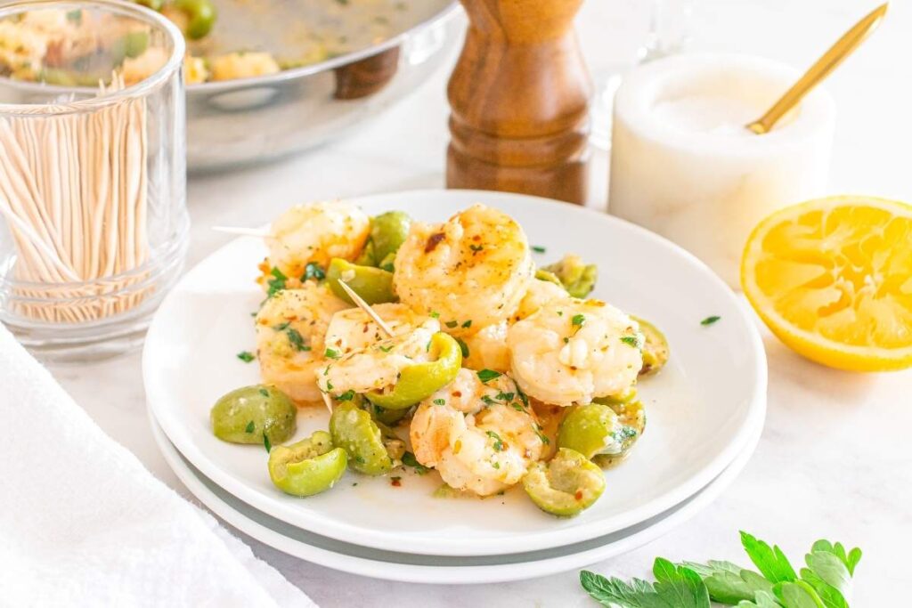 Dirty martini shrimp on a plate, garnished with toothpicks, with lemon, salt, and extra toothpicks in the background.