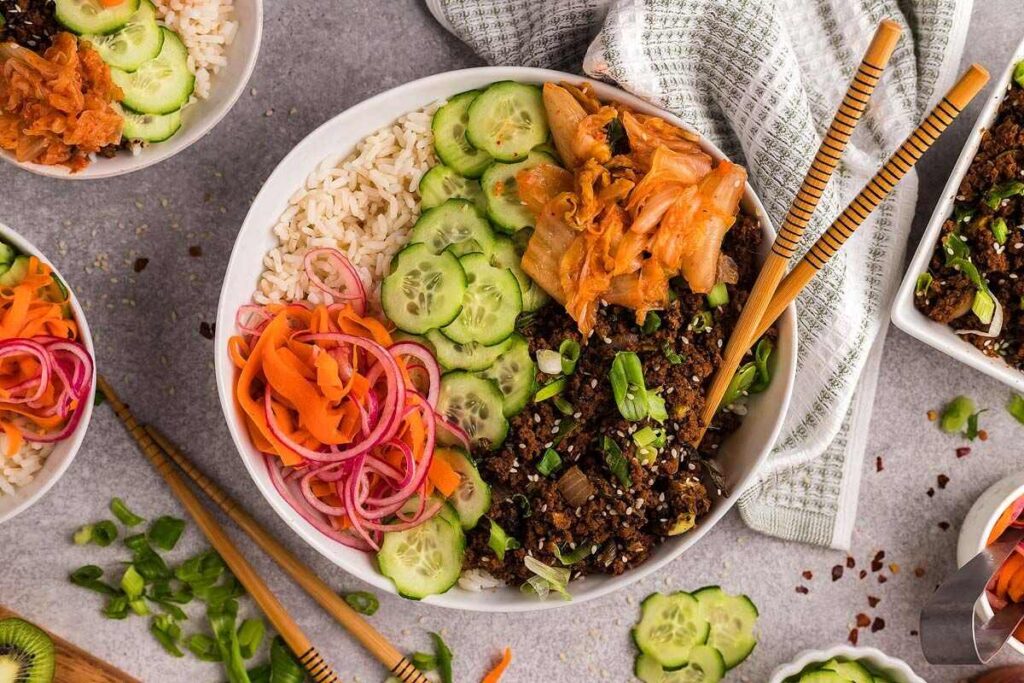 An easy Beef Bulgogi Bowl with rice, ground beef, kimchi, cucumbers, and pickled veggies, served with chopsticks on a gray surface.