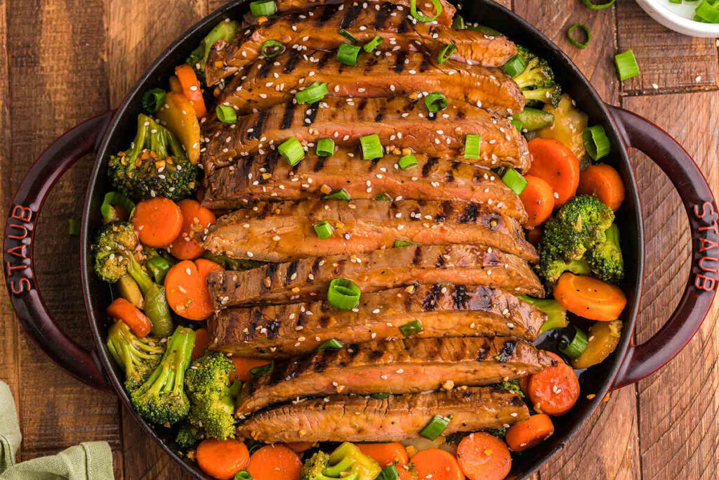 Grilled teriyaki flank steak slices with mixed vegetables, sesame seeds, and green onions in a skillet.