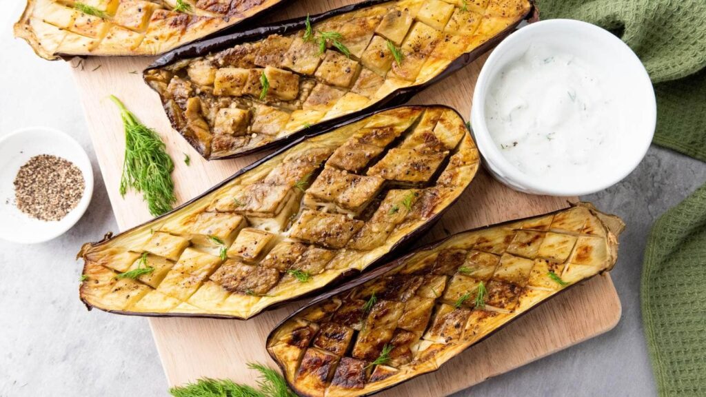 Slices of baked eggplant on a wooden board next to a ramekin of white sauce.