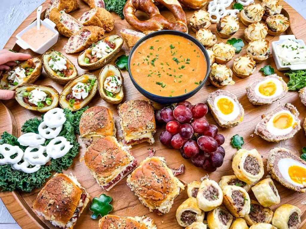 A large wooden platter laden with a variety of appetizers, including stuffed potato skins topped with sour cream, mini sliders, scotch eggs, sausage rolls, pretzel bites, and a bowl of soup, alongside a cluster of red grapes.
