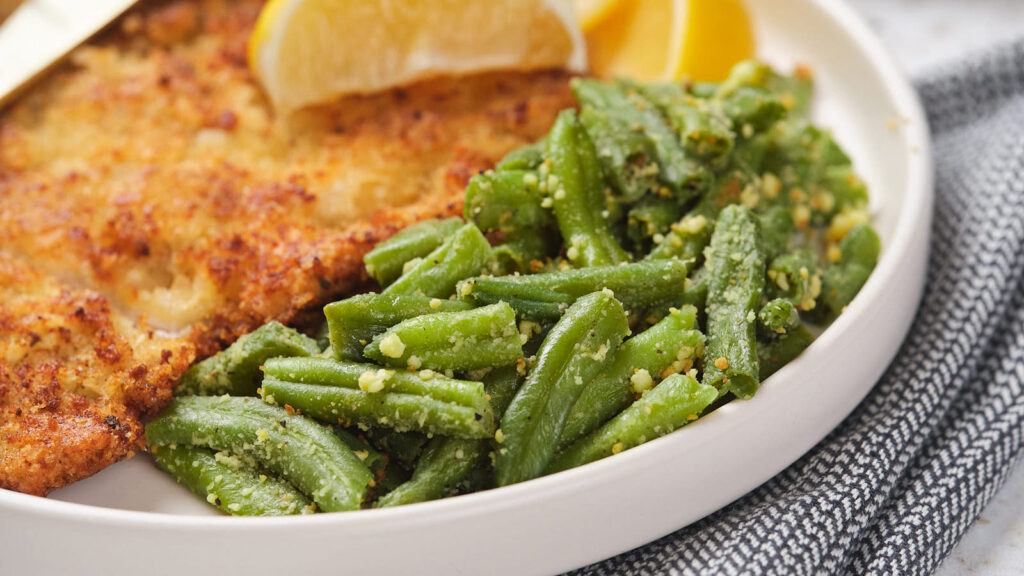 Roasted green beans on a white plate, served with chicken fillet and lemon slices.
