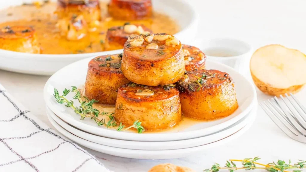 Stacked garlic butter fondant potato rounds with golden-brown edges on a white plate, garnished with herbs and bathed in a buttery sauce.