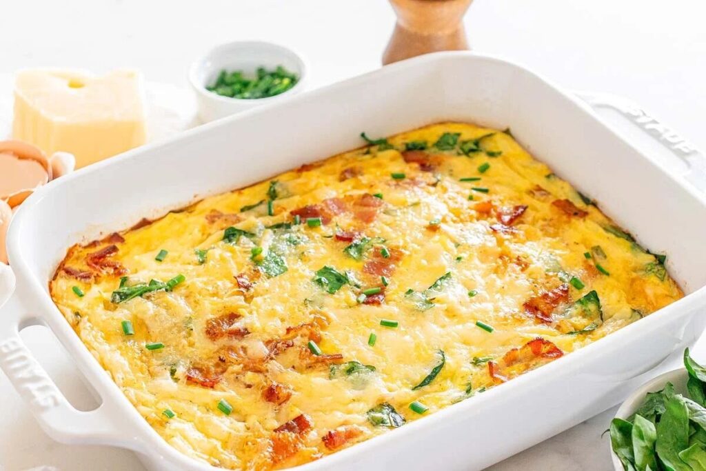 Hashbrown casserole baked in a white dish and garnished with fresh herbs. Eggs, cheese, chives, and a pepper grinder sit nearby.