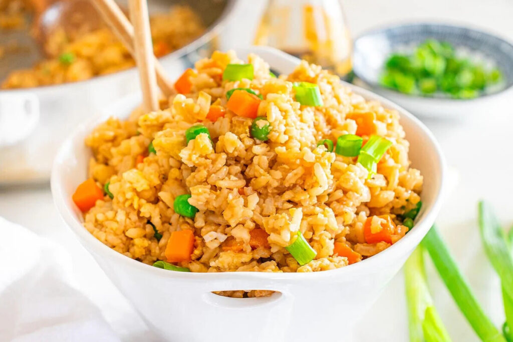 A bowl of hibachi fried rice with peas, carrots, and green onions, accompanied by chopsticks.