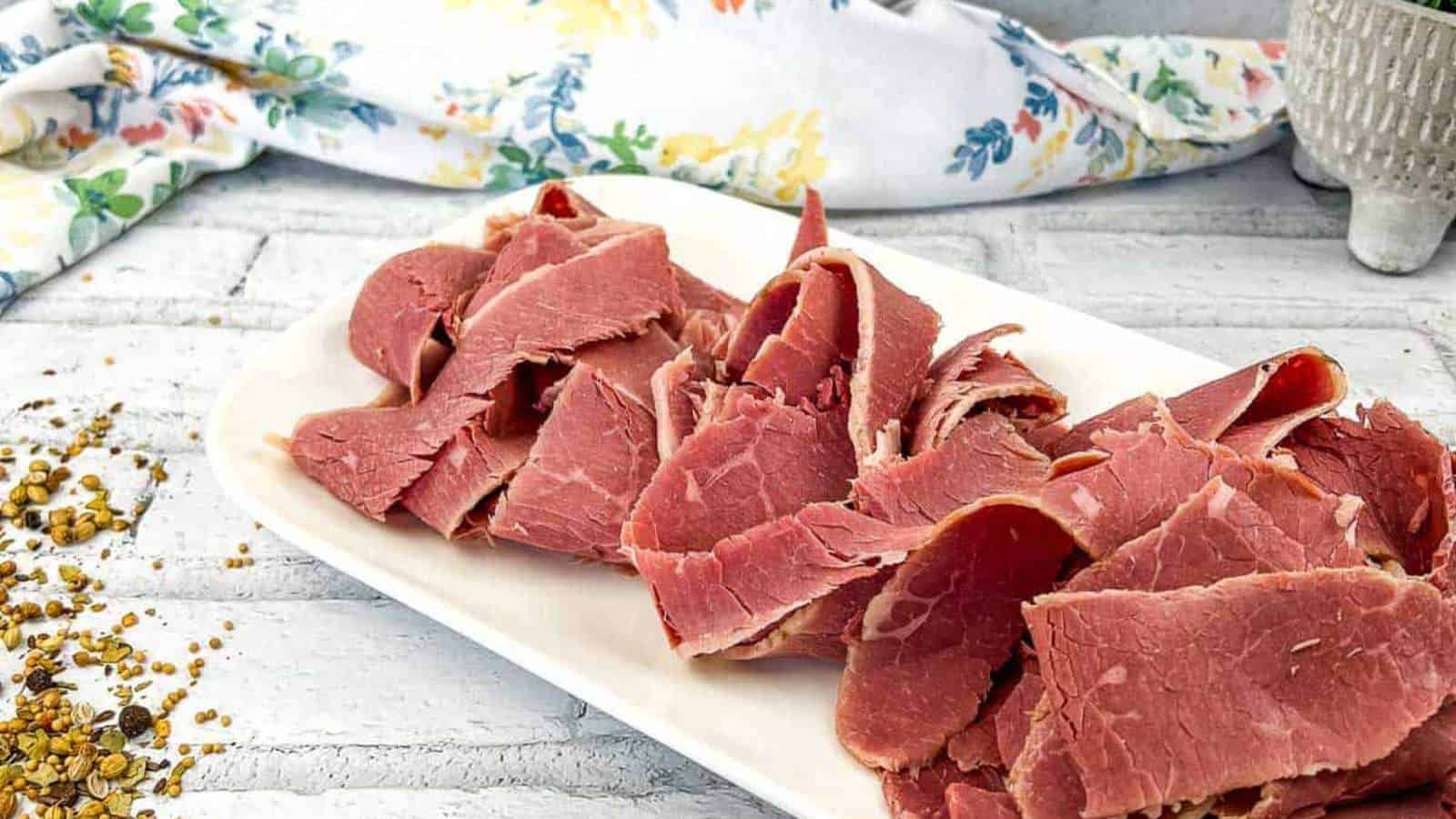 A plate of Homemade Corned Beef on a white rectangular dish, placed on a white brick surface with a floral cloth and seeds nearby.