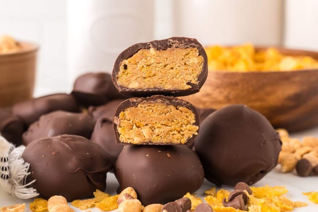A stack of two chocolate-covered peanut butter balls, reminiscent of homemade Butterfinger bites, with a bowl of peanuts and cornflakes in the background.