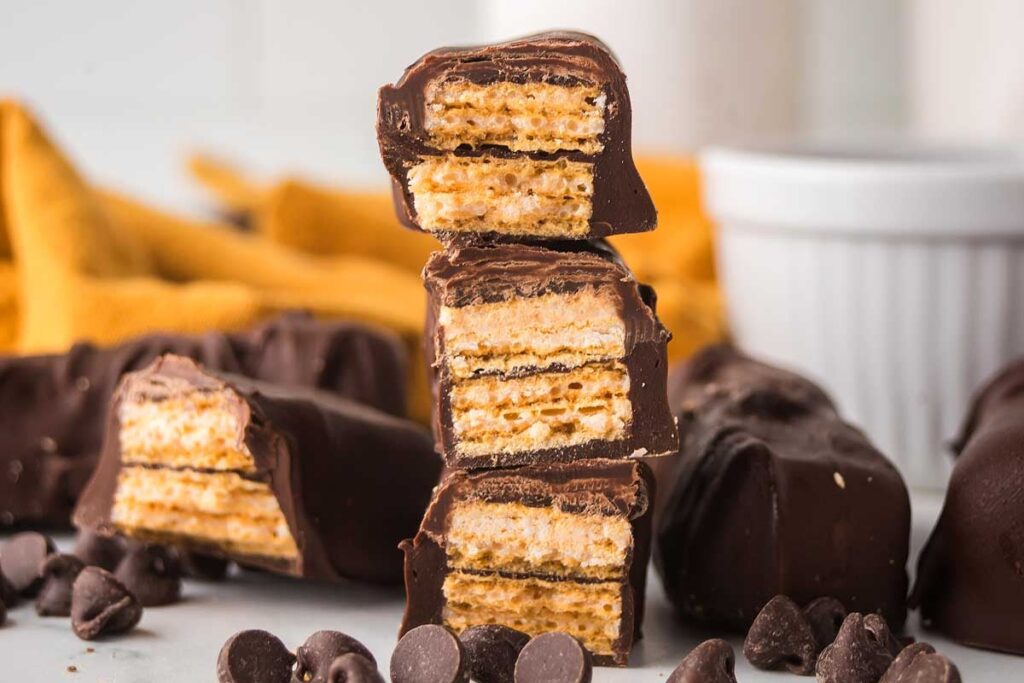 Stacked homemade Kit Kat bars with wafer layers sit among chocolate chips on a yellow cloth.