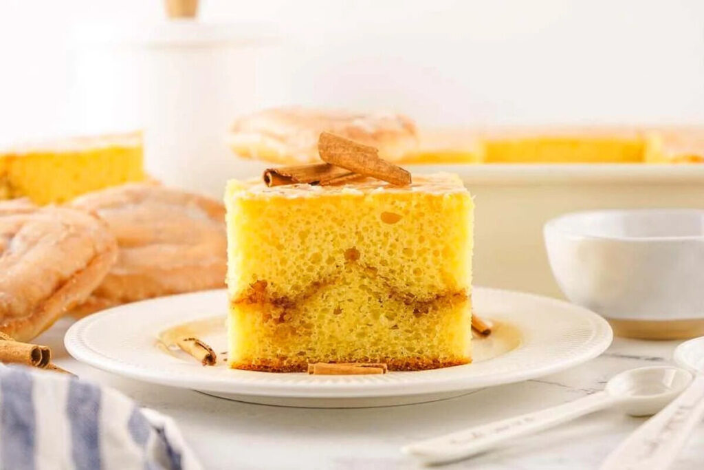 A slice of an irresistible honey bun cake recipe, sits delicately on a plate topped with aromatic cinnamon sticks.