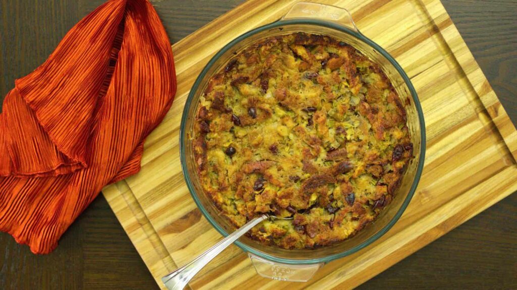A glass baking dish filled with a baked stuffing sits on a wooden cutting board. A serving spoon is in the stuffing, and a red cloth is draped to the left, perfect for cozy fall recipes.