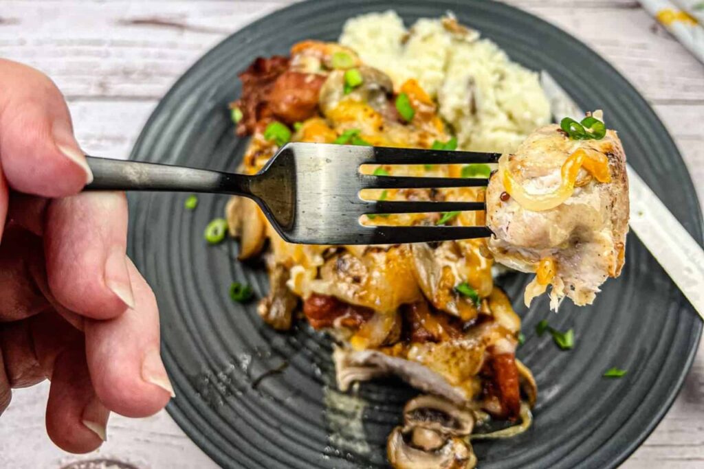 A close-up of a fork holding a bite of chicken topped with cheese and vegetables, with a plate of the same meal and rice in the background.