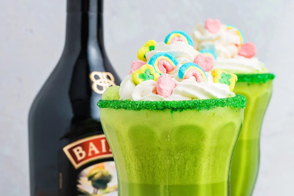 A green Irish cream milkshake topped with whipped cream and colorful cereal sits beside a bottle of Baileys Irish Cream.
