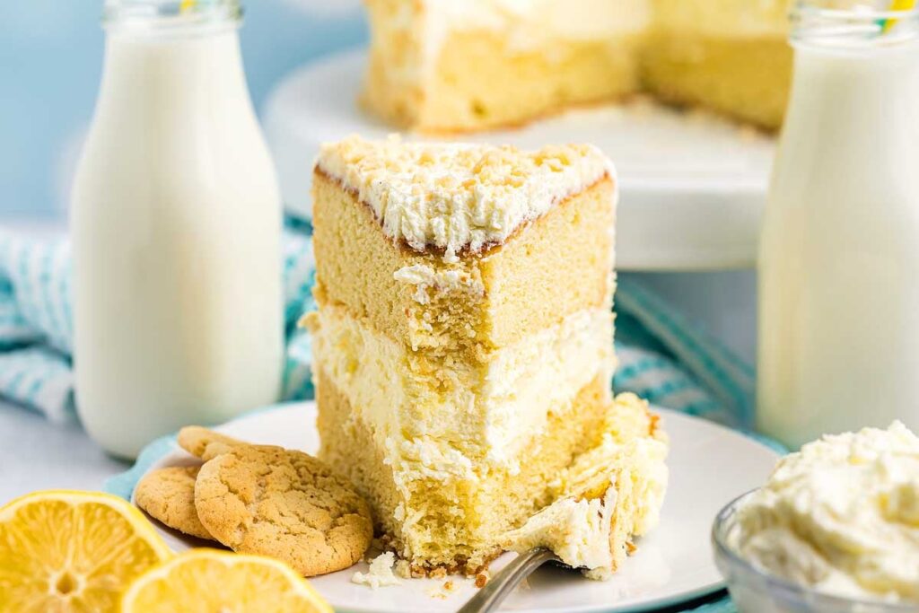 A slice of Italian lemon mascarpone cake with frosting, served with lemon cookies, a fork, and milk, with the rest of the cake in the background.
