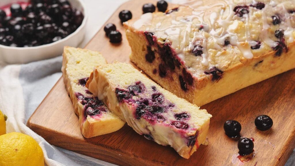 A loaf of blueberry bread on a wooden cutting board, with slices cut to show the interior.