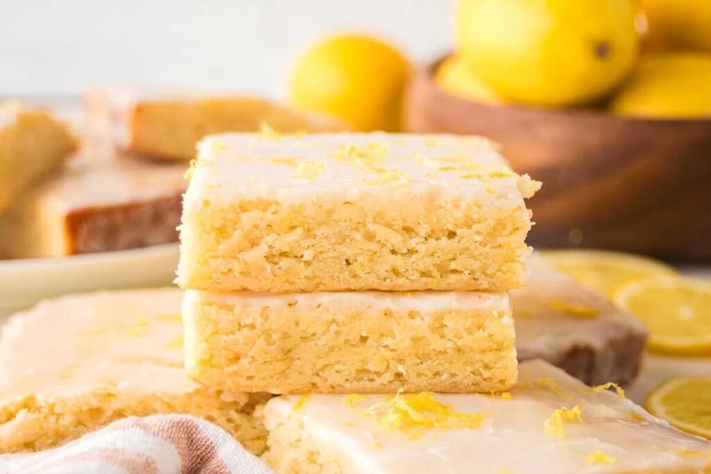 Two stacked lemon brownies with glaze are surrounded by more bars and a bowl of fresh lemons in the background.