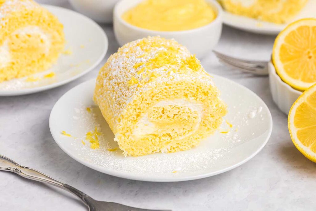 A slice of lemon cake roll with cream filling, dusted with powdered sugar, sits on a white plate.