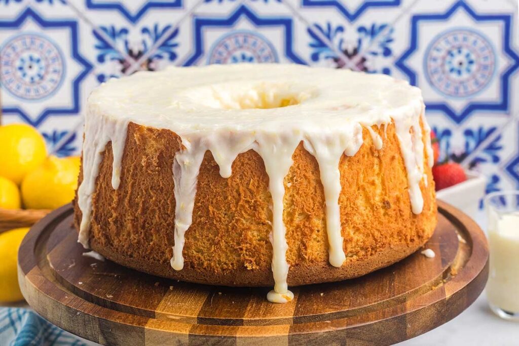 Lemon chiffon cake with white icing, served on a wooden stand with fresh lemons and strawberries.