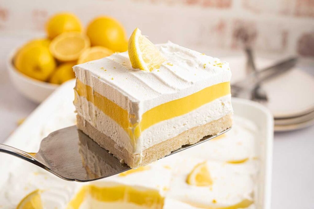 Lemon lush dessert bar with layers and a lemon slice on a spatula.