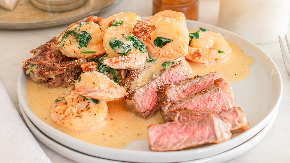 A plate with slices of steak and creamy shrimp in sauce, garnished with spinach.