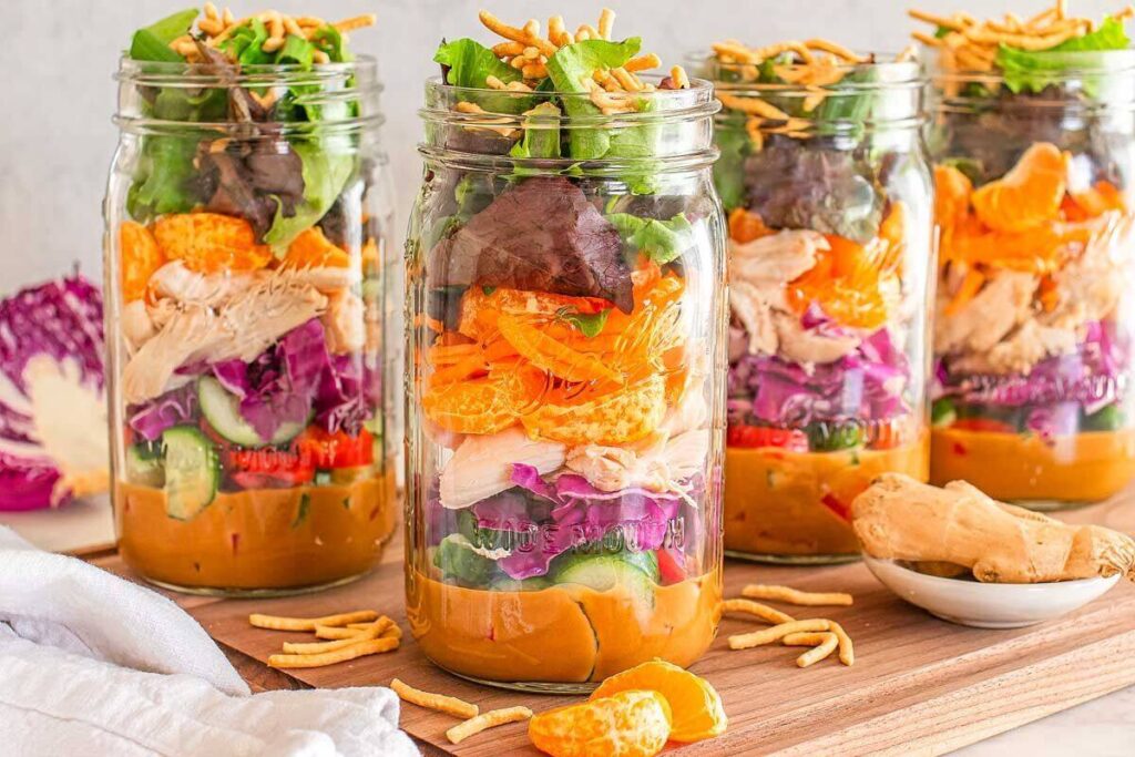 Four Mason jars filled with layered Asian chicken salad ingredients, including lettuce, mandarins, chicken, cucumber, and purple cabbage, are artfully arranged on a wooden board.