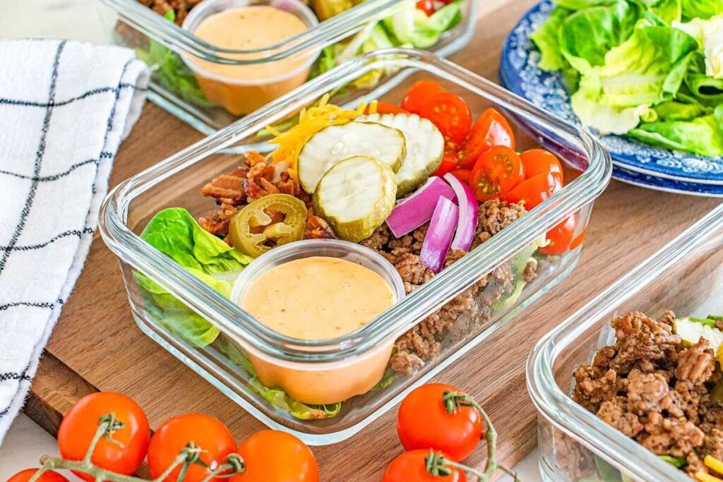 Glass containers with vibrant taco salads featuring ground beef, cherry tomatoes, pickles, onions, jalapeños, cheese, and dressing.