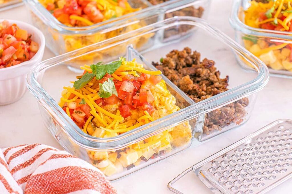 Glass meal prep container with breakfast taco bowls of ground meat, shredded cheese, and diced tomatoes, placed beside a grater.