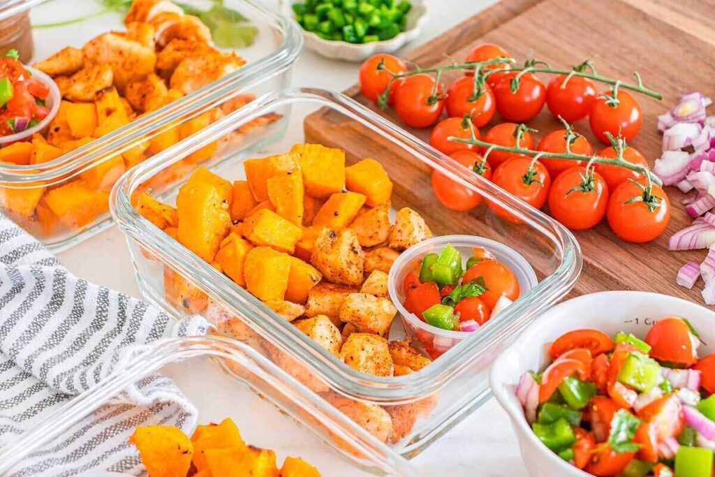 Glass meal prep containers hold Cajun chicken, roasted sweet potatoes, and fresh tomato salsa, making these bowls a colorful kitchen-ready meal.