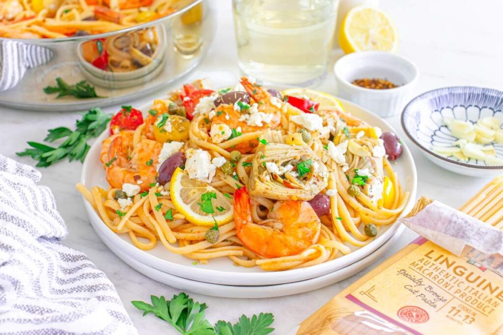 Mediterranean shrimp linguine sits on a white plate, topped with vegetables, herbs, and feta cheese.