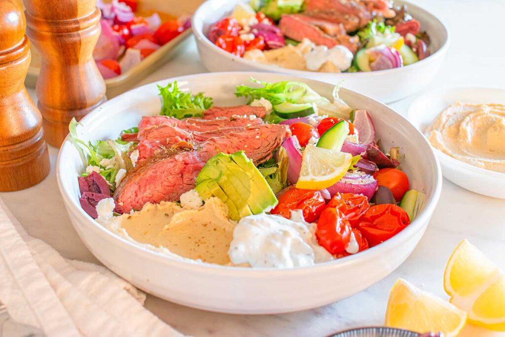 A Mediterranean steak bowl with sliced beef, grilled vegetables, avocado, hummus, tzatziki, and lemon wedges on a marble surface.