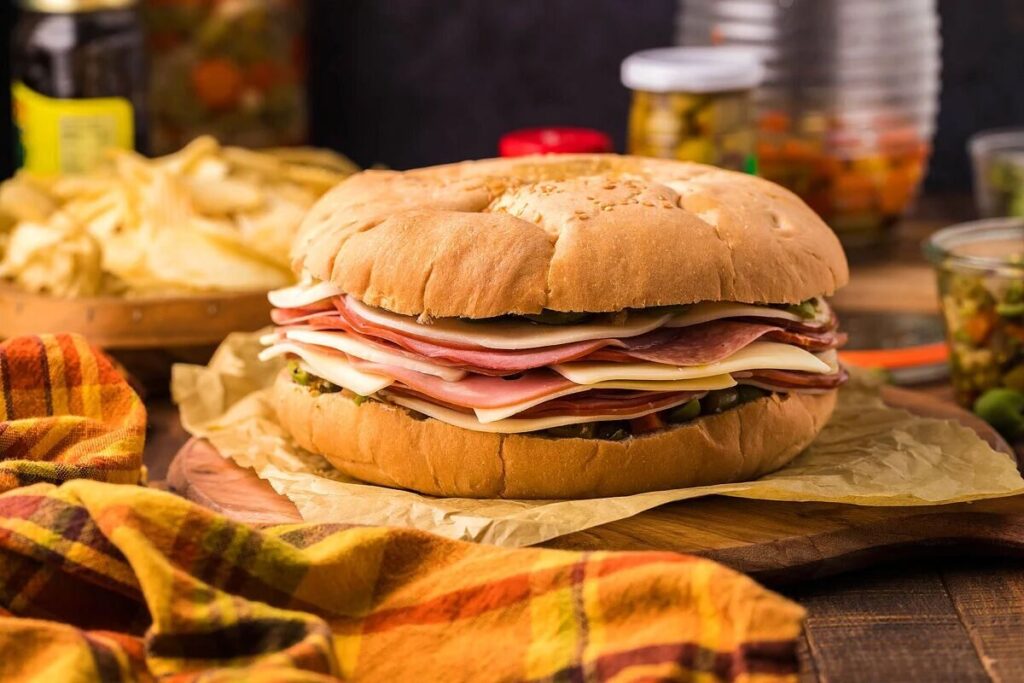 Large muffaletta sandwich with layers of deli meats, cheese, and olive spread on a round bun, surrounded by chips and jars of pickles.