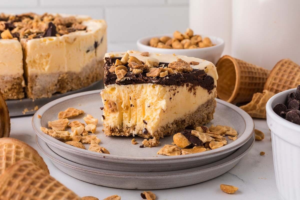 Slice of cake on a plate, topped with chocolate and nuts, surrounded by waffle cones, and bowls of nuts and chocolate chips.