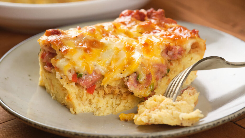 A slice of baked casserole with layers of cheese, ham, and vegetables on a plate, with a fork beside it.