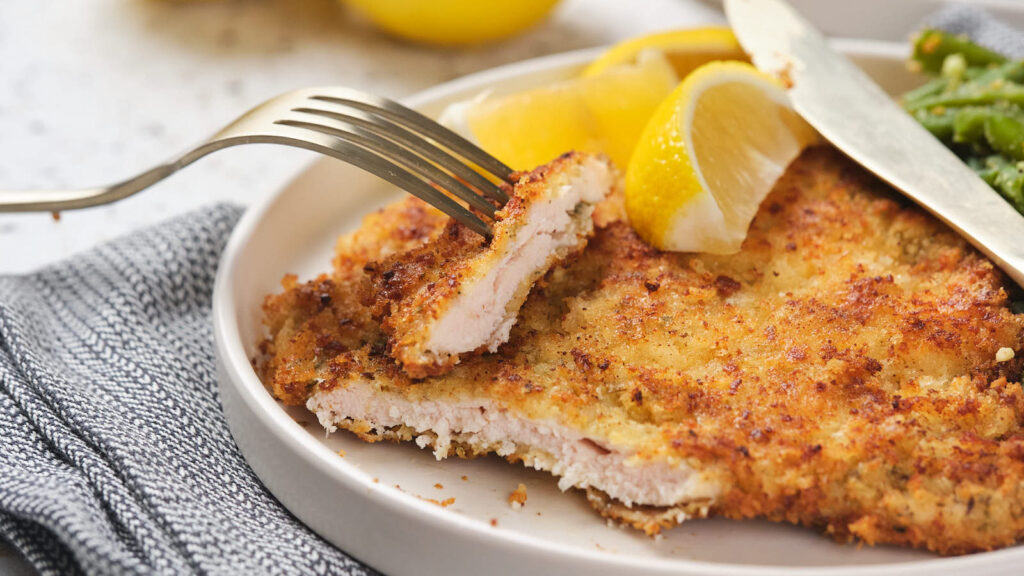 A plate of crispy fried chicken cutlet with a fork, garnished with lemon slices and a side of green beans.