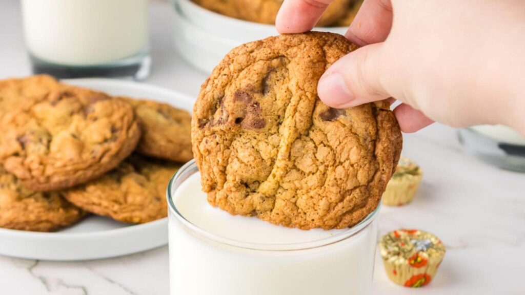 A hand is dipping a chocolate chip cookie into a glass of milk, with more cookies on a plate in the background.