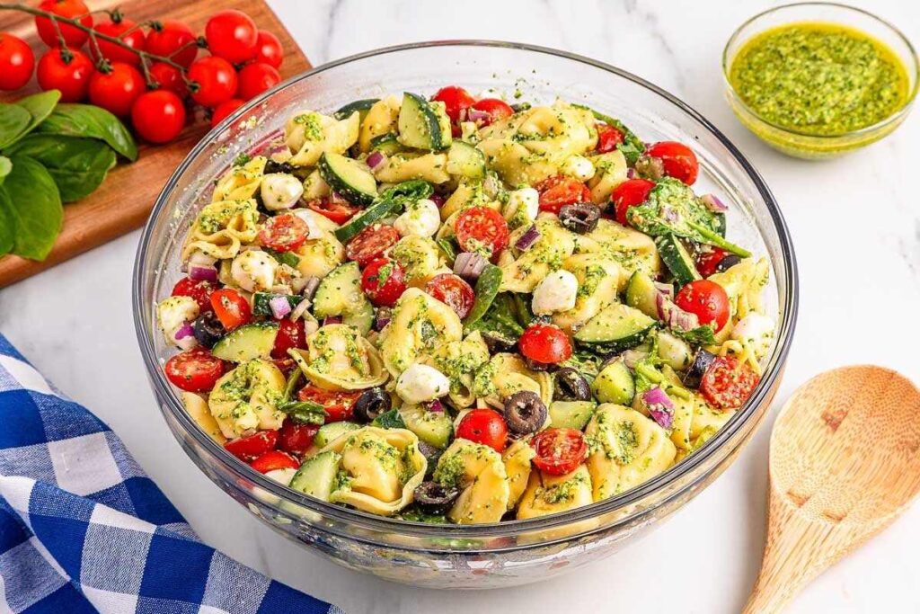 Pesto tortellini pasta salad with tomatoes, cucumber, olives, and mozzarella on a marble surface.