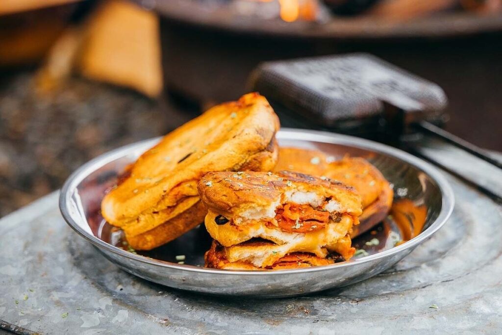 Three pie iron pizzas with melted cheese and pepperoni are toasted to perfection on a metal plate, one partially eaten.