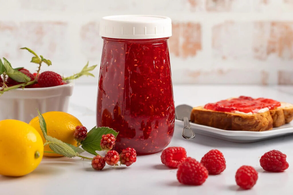 A glass jar of vibrant red raspberry jam sits on a white surface.