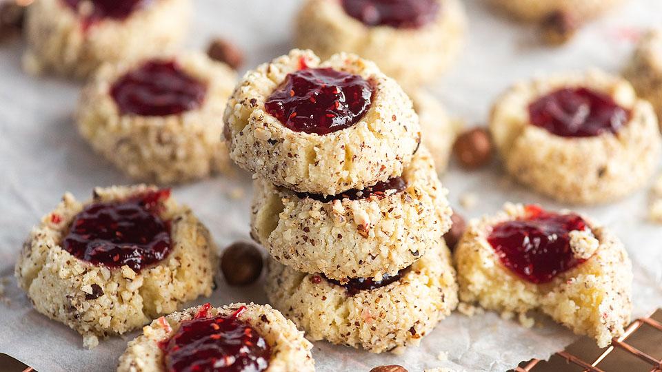 Cookies with red jam centers and finely chopped nuts on a parchment-lined surface.