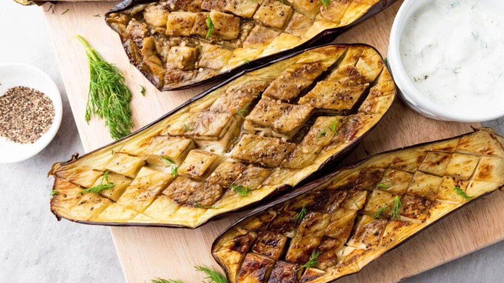 Grilled or roasted eggplants sliced in half, arranged on a wooden cutting board with a bowl of yogurt sauce.