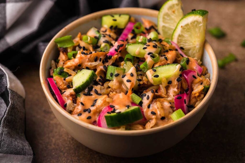 A spicy tuna rice bowl with rice, avocado, edamame, tuna, and cucumber, topped with sauces and toasted crumbs, served with chopsticks.