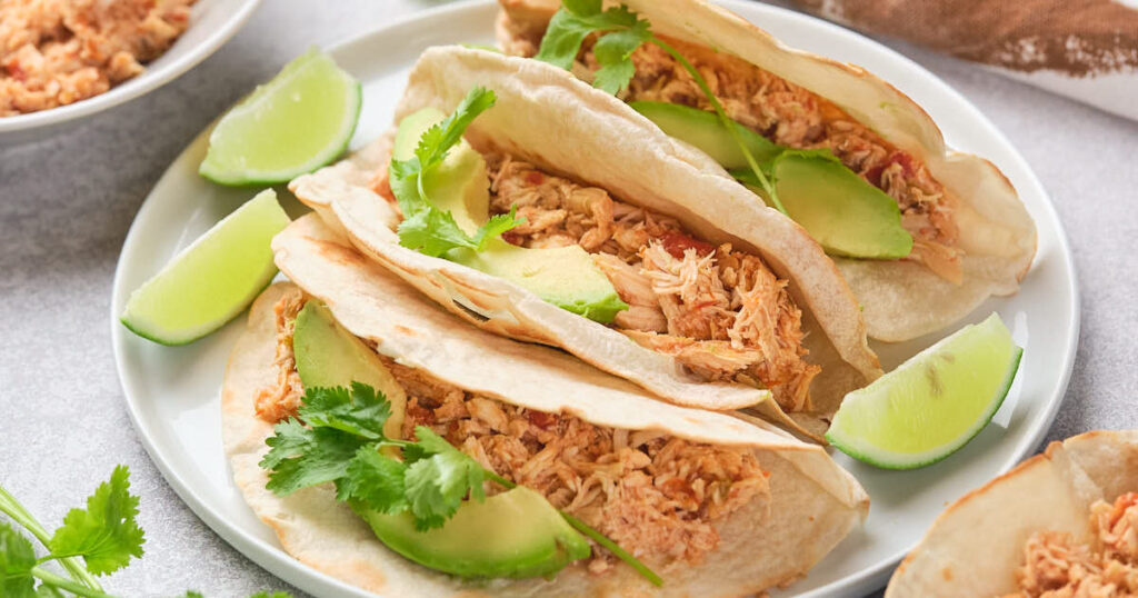Three tacos filled with shredded chicken, garnished with cilantro and lime wedges, served on a white plate.