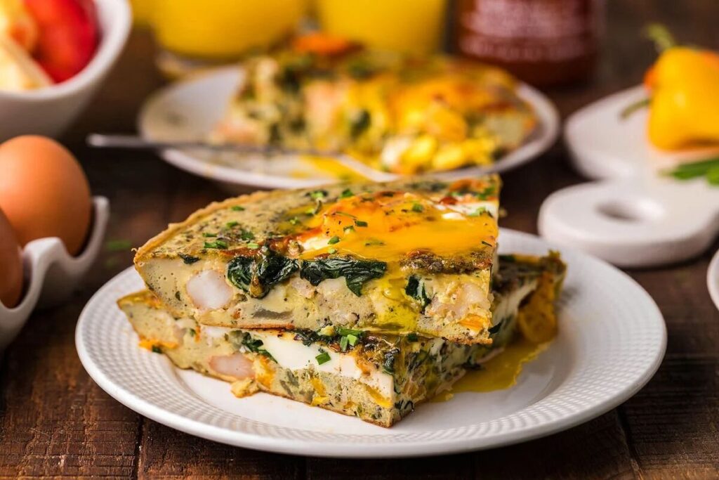 Two slices of vegetable frittata on a white plate with eggs and fresh vegetables in the background.