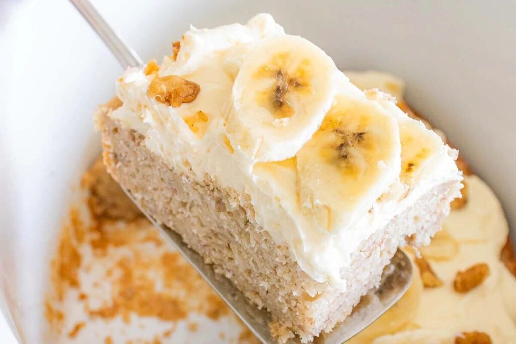 A slice of slow cooker banana cake topped with frosting and banana slices is being lifted from the baking dish with a spatula.