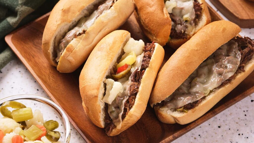A wooden platter with several sandwich rolls filled with beef, pickles, and other condiments.