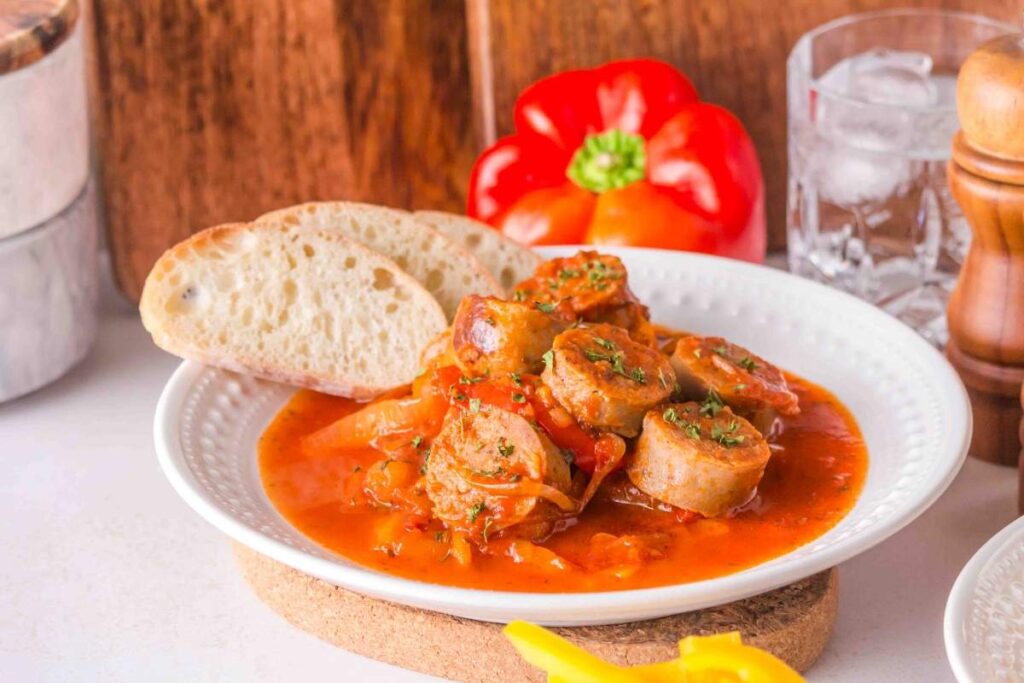 Plate of slow cooker sausage and peppers in tomato sauce with herbs, served with a slice of bread and a red bell pepper in the background.