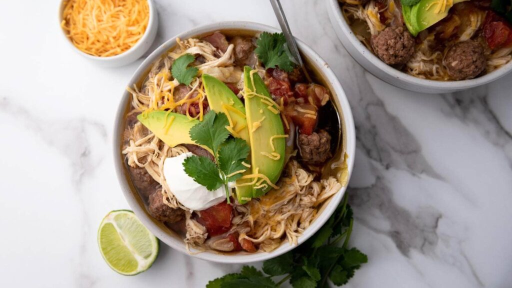 A bowl of chicken and beef soup topped with avocado slices, cilantro, sour cream, and shredded cheese, next to a lime wedge.