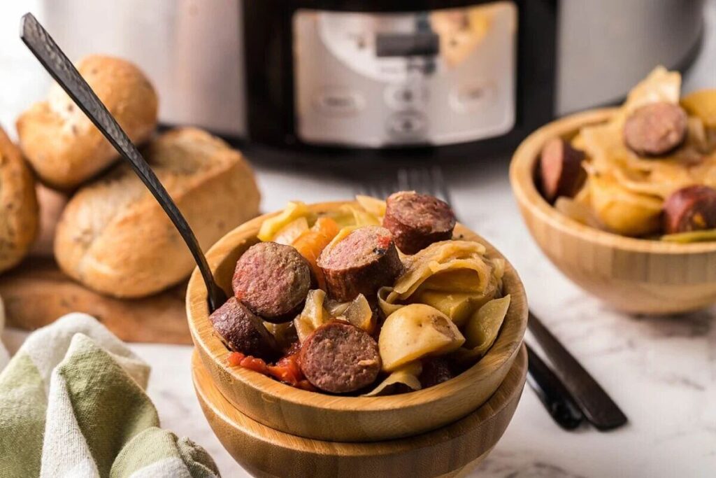 A wooden bowl of slow cooked cabbage, potatoes, and kielbasa bratwurst or sausage with bread rolls, with a slow cooker in the background.